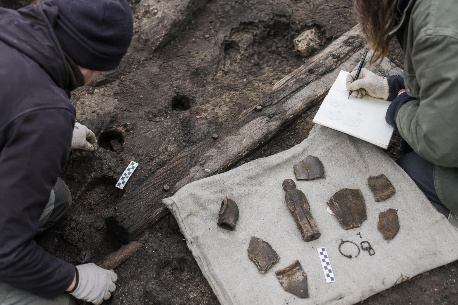 Dwoje archeologów przy odkrywce, na płótnie ułożone drewniane i ceramiczne zabytki
